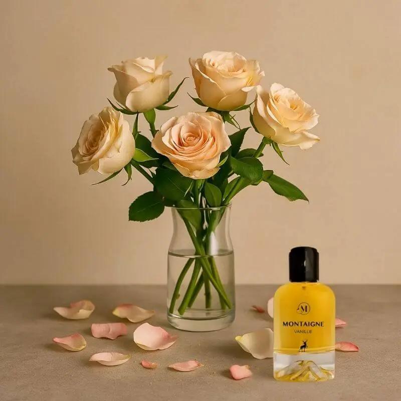 Parfum Montaigne Vanille en flacon jaune, posé sur une table avec des pétales de rose, à côté d’un vase de roses pâles